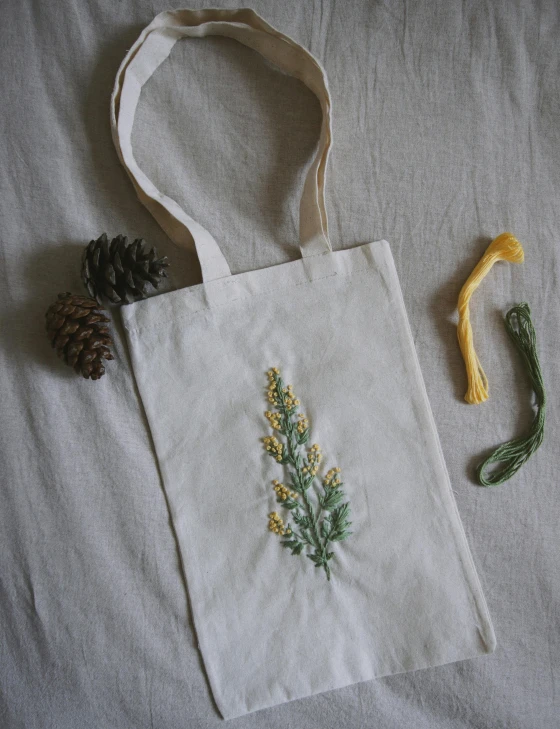 a tote bag sitting on top of a bed next to a pine cone, a cross stitch, by Dóra Keresztes, wilted flowers, product view, marigold, herbarium page