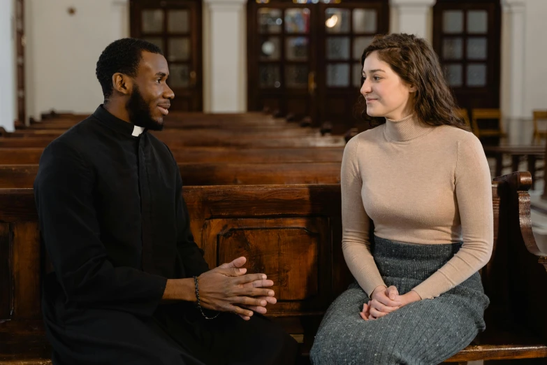 a man and a woman sitting on a bench in a church, dark people discussing, he is wearing a brown sweater, priest, black man