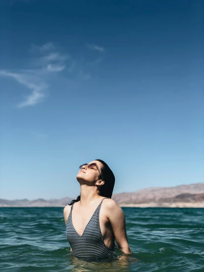 a woman standing in the middle of a body of water, in the sun, looking upwards, striped, looking to the side off camera