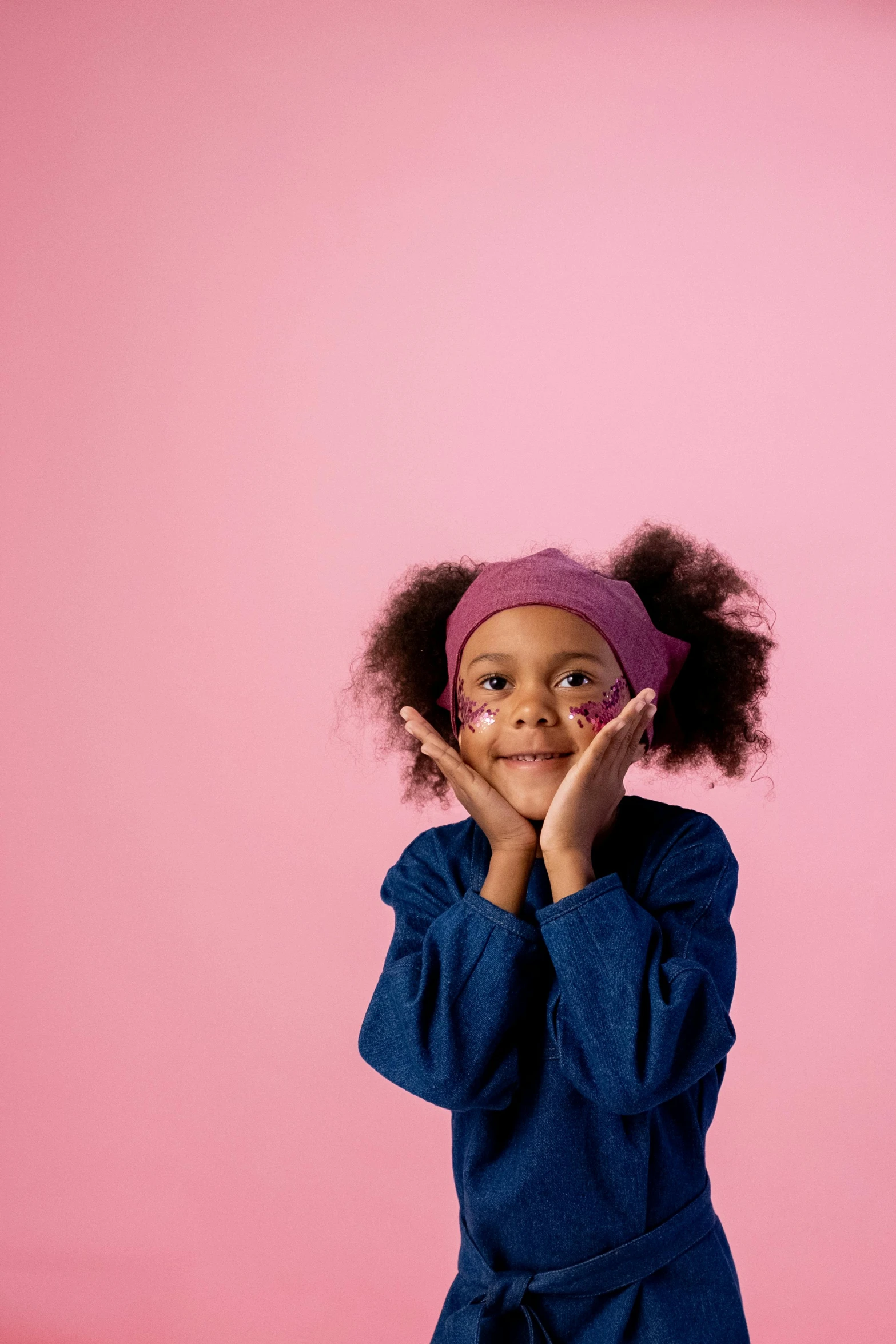 a little girl standing in front of a pink wall, hands on face, wearing a headband, ( ( dark skin ) ), indigo