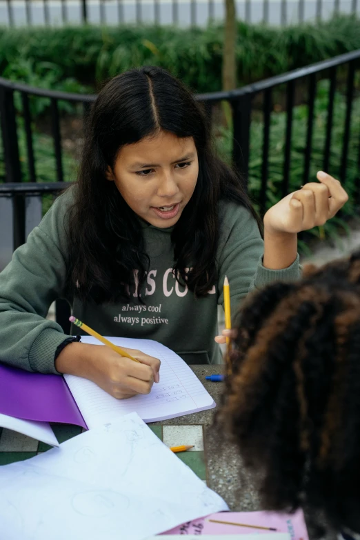 two girls sitting at a table with papers and pencils, by Loren Munk, pexels contest winner, alexandria ocasio - cortez, outdoor photo, teaching, determined expression