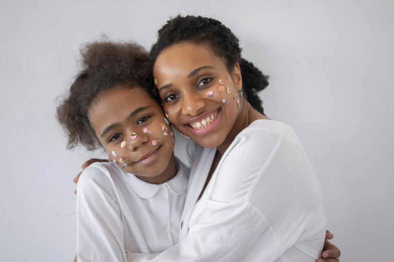 a woman and a young girl hugging each other, by Lily Delissa Joseph, pexels contest winner, markings on her face, on white background, face accessories, glow up