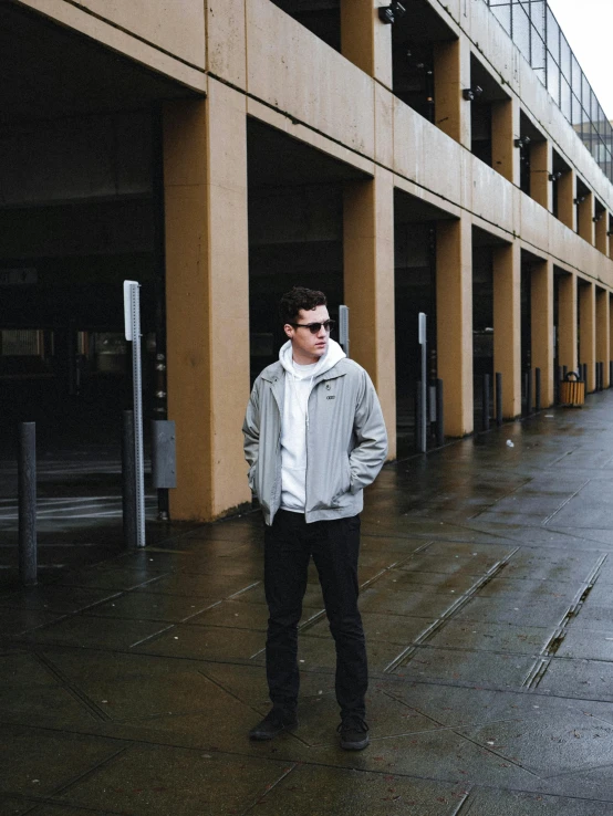 a man standing on a sidewalk in front of a building, inspired by Jean Malouel, unsplash, grey hoodie, wearing an aviator jacket, slightly pixelated, pale grey skin