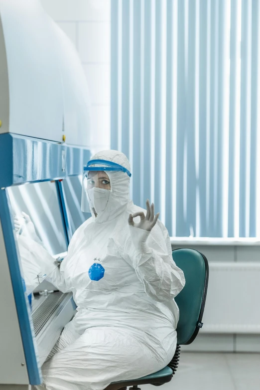 a man in a medical gown sitting in a chair, by Adam Marczyński, shutterstock, private press, an astronaut giving a peace sign, hazmat suits, working in her science lab, photograph taken in 2 0 2 0