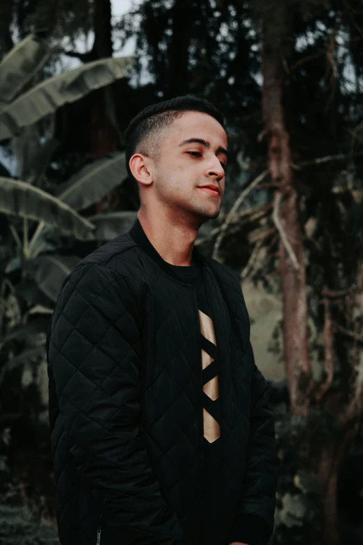 a man standing in front of a lush green forest, an album cover, by Robbie Trevino, pexels contest winner, wearing a black sweater, patterned clothing, headshot profile picture, slightly tanned