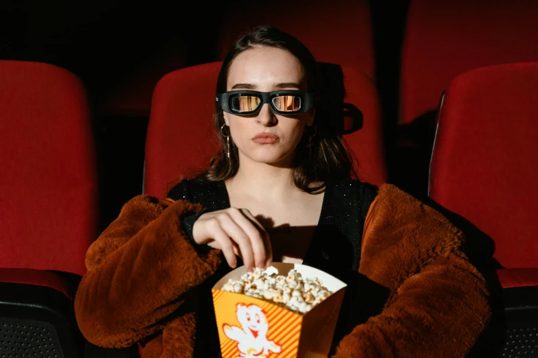 a woman sitting in a chair with a box of popcorn, inspired by Elsa Bleda, trending on pexels, digital glasses, dilraba dilmurat, in a cinema, wearing orange sunglasses