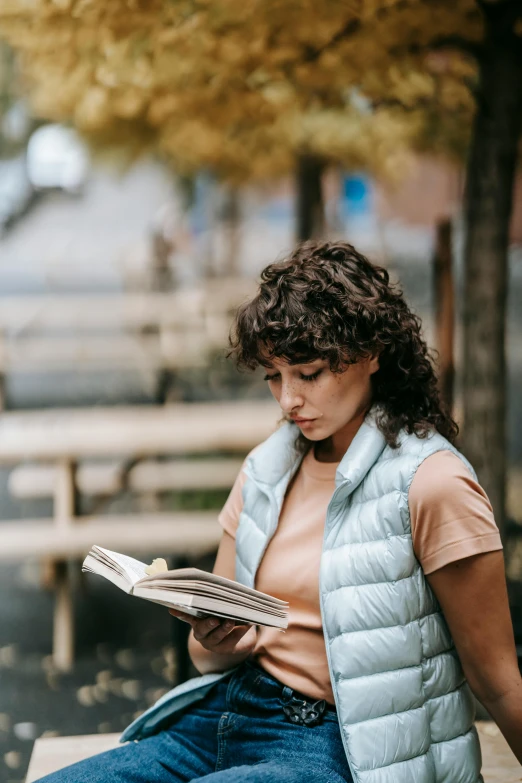 a woman sitting on a bench reading a book, a portrait, pexels contest winner, happening, curly haired, gif, reading engineering book, model wears a puffer jacket