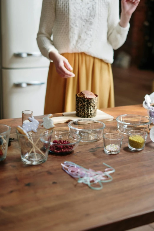 a woman standing in front of a table with a cake on it, process art, glass labware, seeds, cosy, ingredients on the table