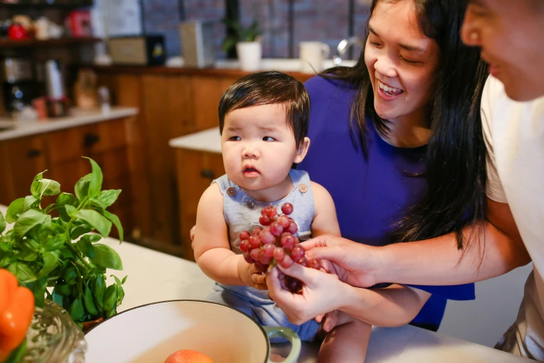 a woman holding a baby next to a bowl of fruit, tyler edlin and natasha tan, vine, portrait image