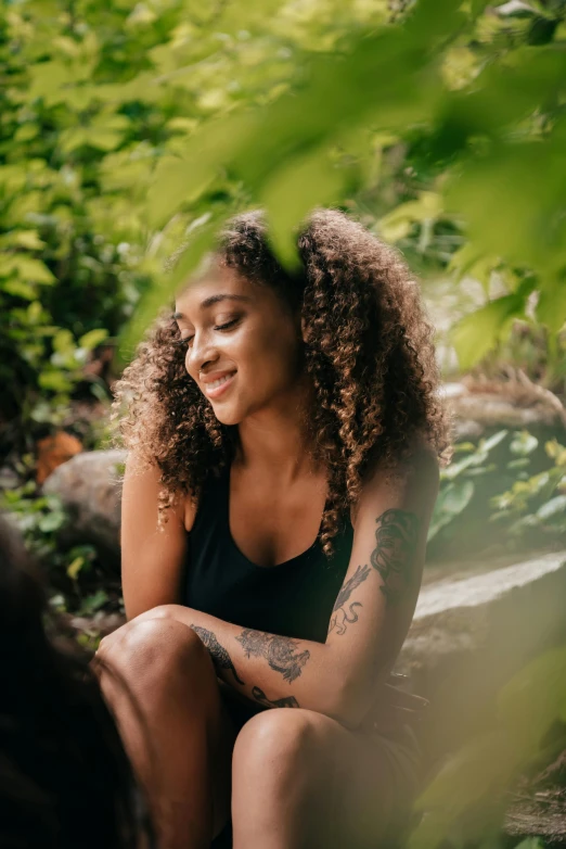 a woman sitting on a rock in the woods, a portrait, pexels contest winner, renaissance, dark short curly hair smiling, ashteroth, with tattoos, lush greens