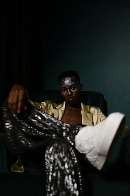 a man sitting in a chair in a dark room, an album cover, by Jessie Alexandra Dick, trending on pexels, afrofuturism, yoruba body paint, shiny metallic glossy skin, androgynous person, brown clothes
