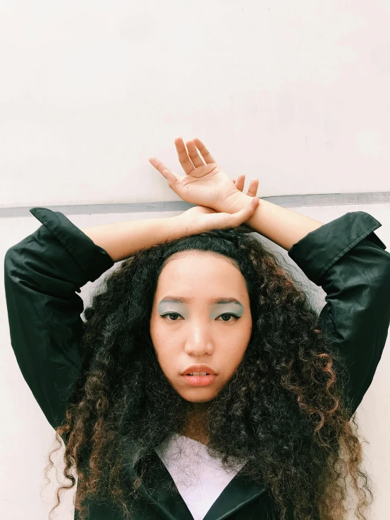 a woman standing in front of a white wall with her hands on her head, an album cover, inspired by Ren Hang, unsplash, long brown puffy curly hair, mixed-race woman, portrait of maci holloway, with index finger