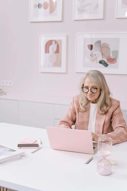 a woman sitting at a table with a laptop, inspired by Alexander Roslin, trending on pexels, arbeitsrat für kunst, wearing a light - pink suit, older woman, gif, in white room