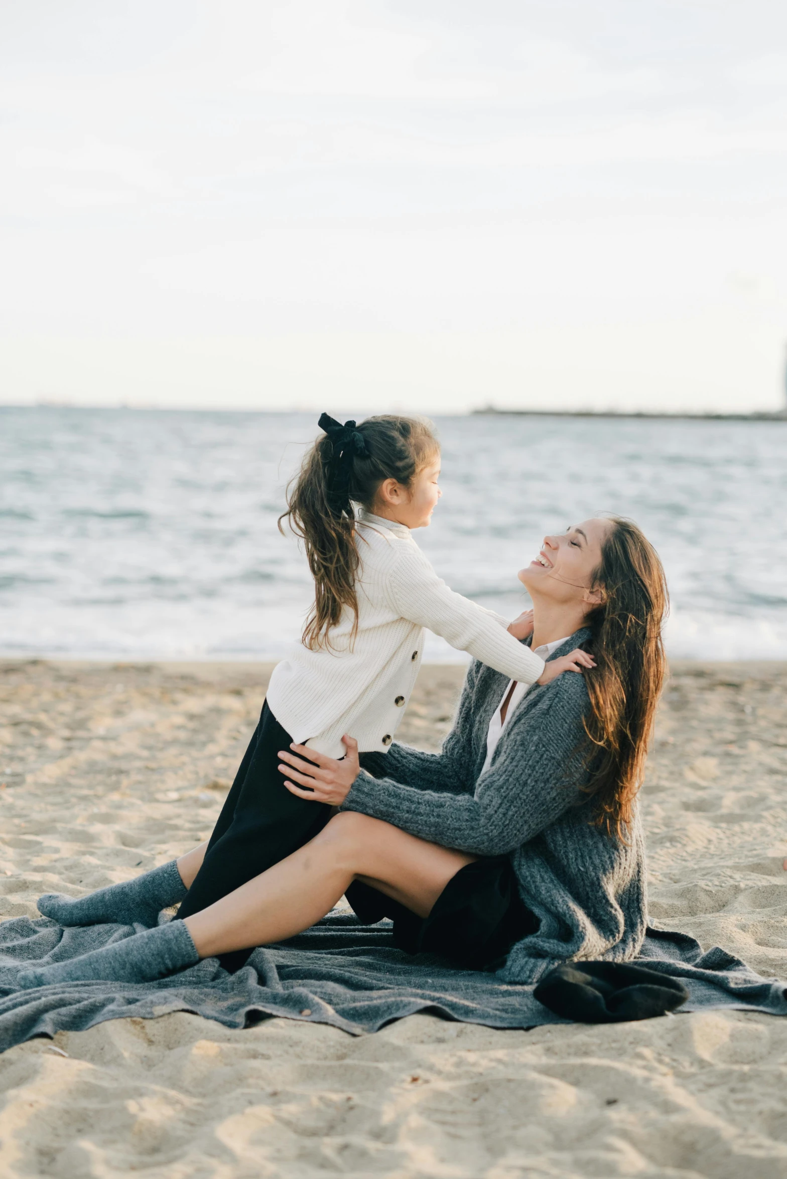 a woman sitting on top of a beach next to a little girl, maternity feeling, portrait featured on unsplash, playful pose, grey