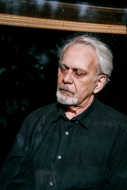 a man in a black shirt sitting in front of a window, an album cover, inspired by Lajos Vajda, slavoj zizek, in front of a black background, solemn expression, sam gilliam