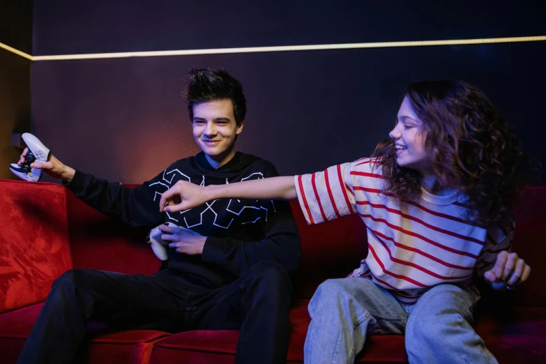 a man and a woman sitting on a couch playing a video game, an album cover, by Adam Marczyński, pexels, antipodeans, joe keery, pointing at the camera, kids playing, in a nightclub