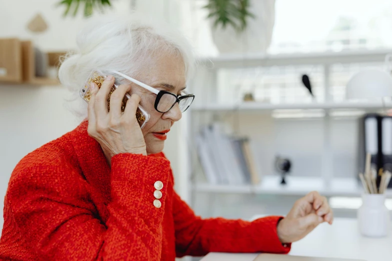 a woman sitting at a desk talking on a cell phone, by Lee Loughridge, trending on pexels, an old lady with red skin, royal commission, white reading glasses, fashionable