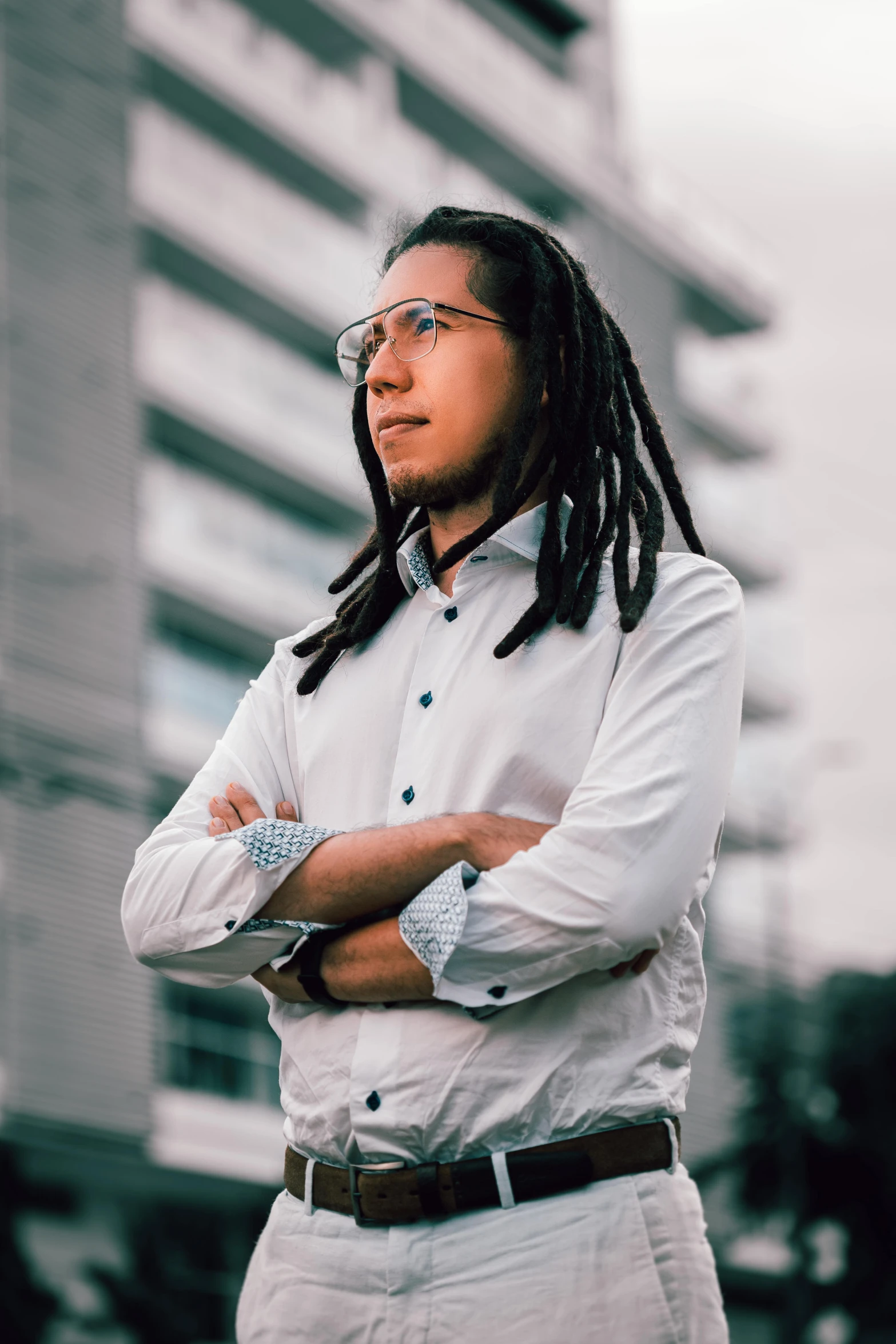 a man with dreadlocks standing in front of a tall building, by Alejandro Obregón, wearing a white button up shirt, avatar image, high quality photo, profile pic