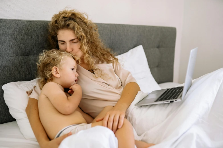 a woman sitting on top of a bed next to a baby, a cartoon, pexels, happening, using a macbook, hugging each other, white, 1x