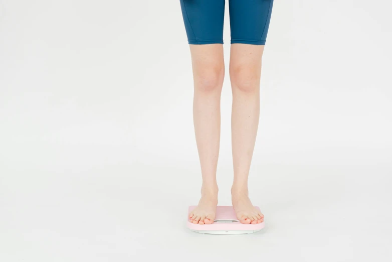 a woman standing on top of a bathroom scale, by Emma Andijewska, pink body, plain background, aoi ogata, knee