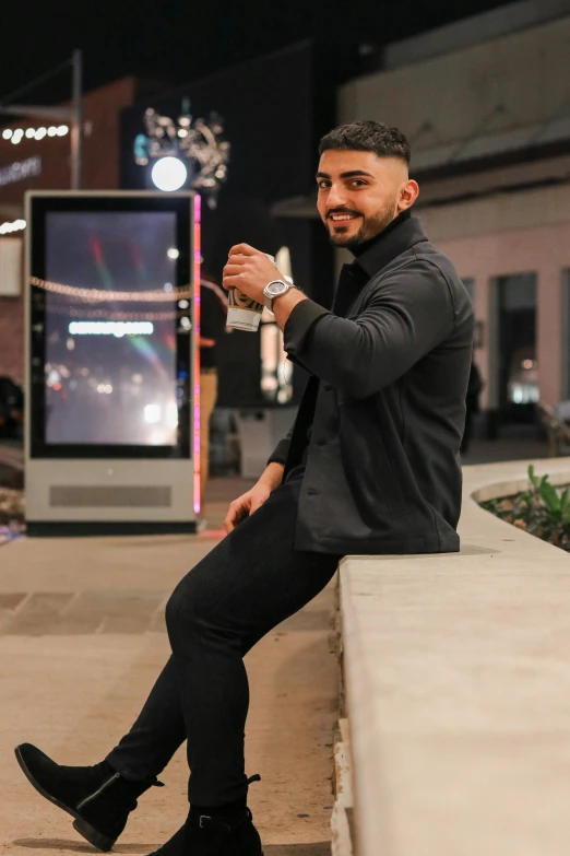 a man sitting on a ledge holding a cup of coffee, kyza saleem, neon lights in the background, posing for a picture, wearing a fancy black jacket