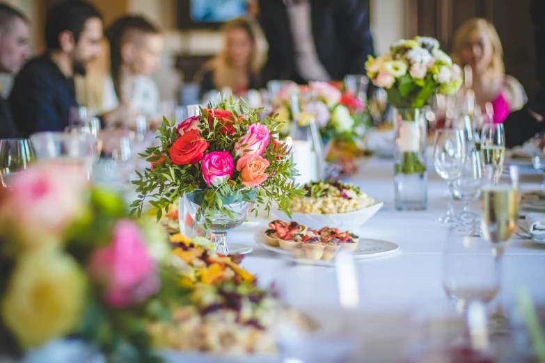 a group of people sitting at a table with plates of food, by Alice Mason, pexels, colourful flowers, champagne on the table, walking down, closeup - view