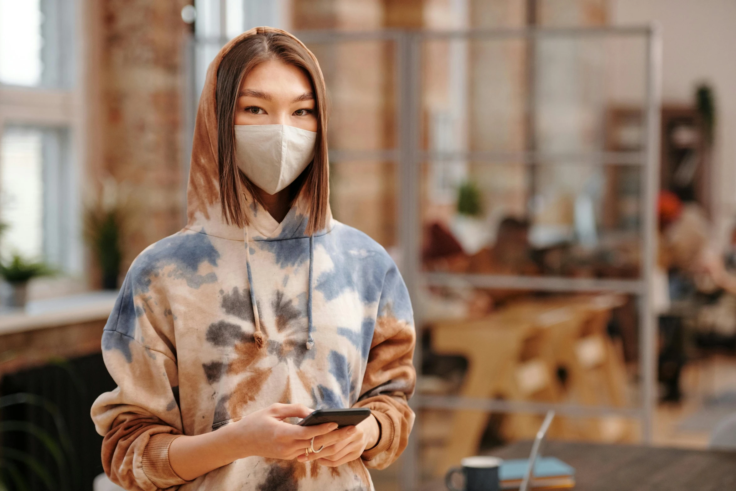 a woman wearing a face mask while looking at her phone, by Aya Goda, trending on pexels, hyperrealism, wearing a tie-dye shirt, wearing a brown, technology cloak, standing in a restaurant