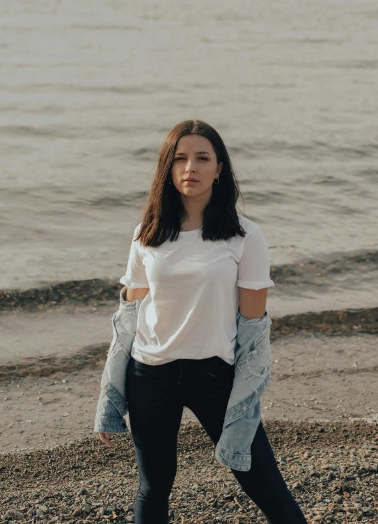 a woman standing on a beach next to the ocean, by Adam Marczyński, white tshirt, young woman with long dark hair, promo image, pokimane