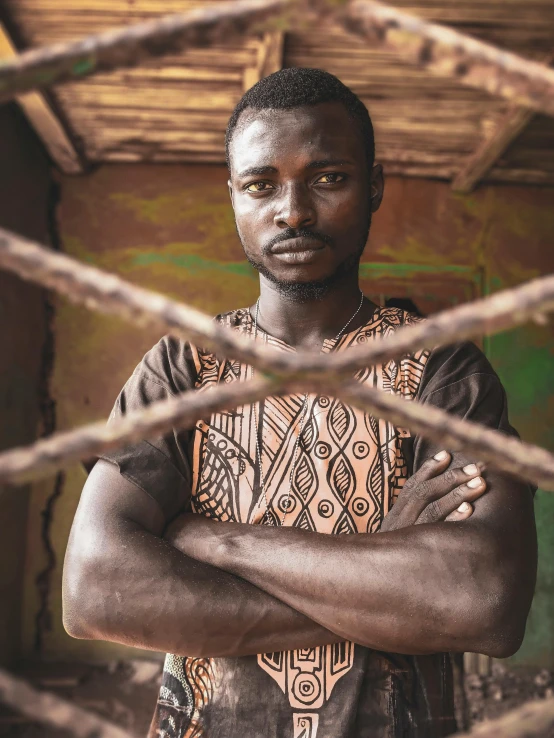 a man standing behind a fence with his arms crossed, by Benjamin Block, pexels contest winner, afrofuturism, wearing an african dress, portrait close up of guy, rustic, stood in a cell