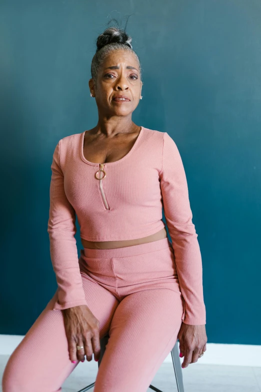 a woman sitting on a chair in front of a blue wall, pink clothes, atiba jefferson, wearing a track suit, muted colored bodysuit