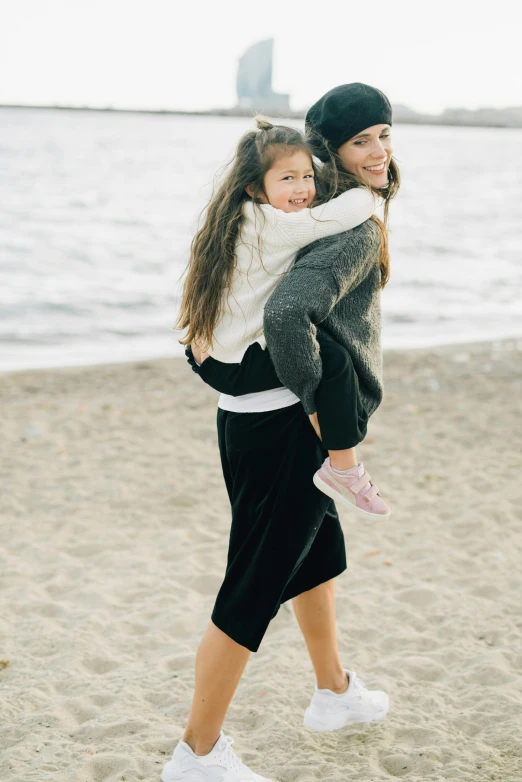 a woman carrying a little girl on the beach, wearing a sweater, stylish pose, forward facing, black