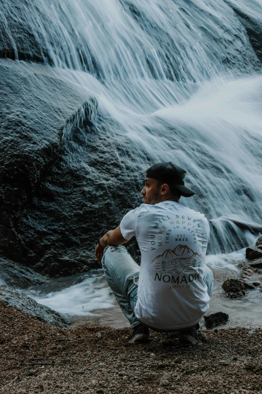 a man sitting in front of a waterfall, inspired by Michael Komarck, unsplash contest winner, wearing a tee shirt and combats, graphic tees, nomad, profile pic