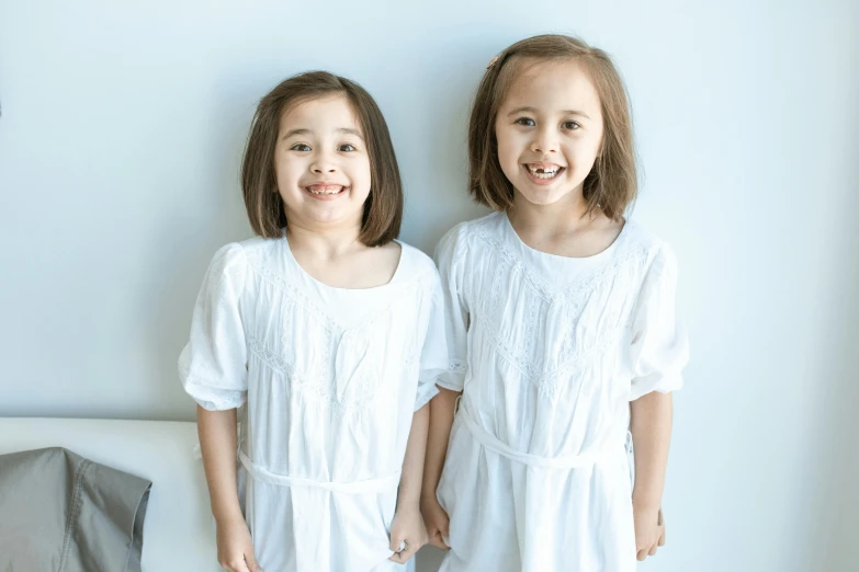 two little girls standing next to each other, inspired by Kate Greenaway, pexels contest winner, antipodeans, wearing a white hospital gown, deep dimples, brunettes, large front teeth