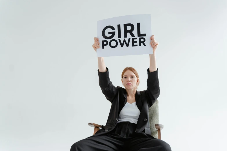 a woman sitting on a chair holding a sign, inspired by Harriet Powers, trending on pexels, feminist art, power girl, a person standing in front of a, profile image, girl with brown hair