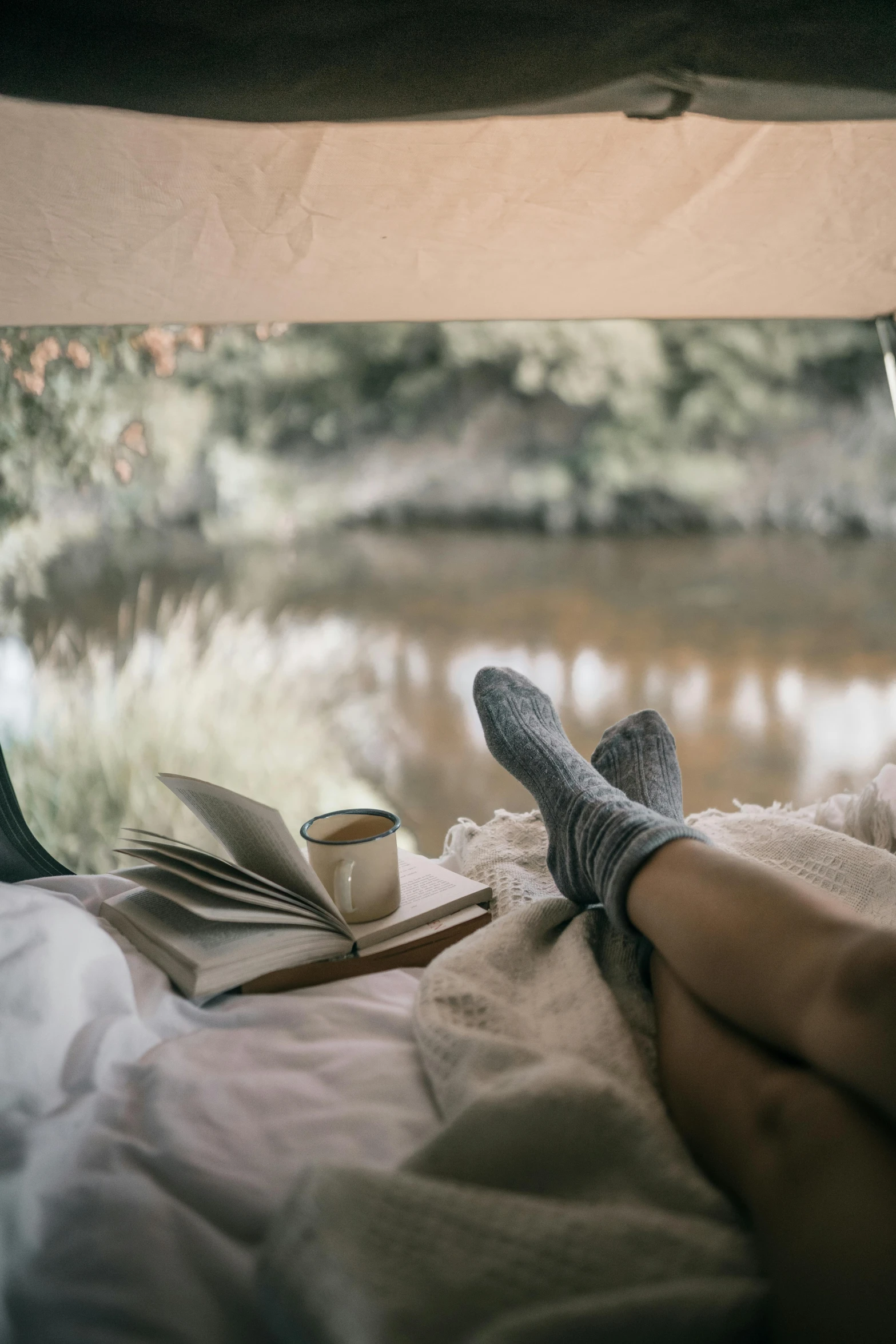 a person laying in a bed with a book and a cup of coffee, by Jessie Algie, unsplash contest winner, near a river, manuka, barefeet, cottagecore