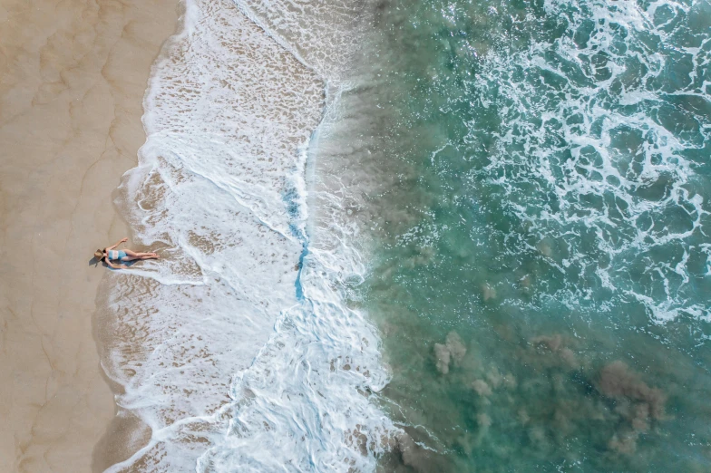 a couple of people laying on top of a sandy beach, by Robbie Trevino, pexels contest winner, cresting waves and seafoam, bird view, woman on the beach, flying over the ocean