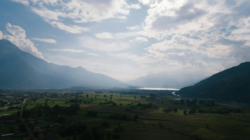 a view of a valley with mountains in the background, a picture, unsplash contest winner, wide river and lake, clouds and fields in background, overhead sun, cinematic footage