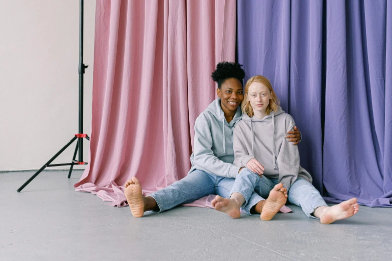 a couple of women sitting next to each other, trending on pexels, visual art, studio backdrop, wearing a hoodie and sweatpants, pastels, with a kid