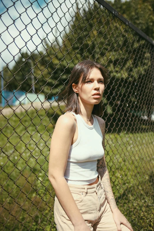 a woman standing in front of a chain link fence, an album cover, inspired by Elsa Bleda, wearing tank top, looking this way, high quality photo, woman with porcelain skin