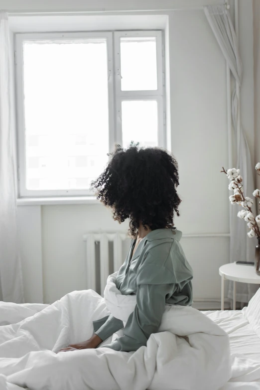 a woman sitting on top of a bed next to a window, trending on pexels, black curly hair, heartbreak, pastel', back view also