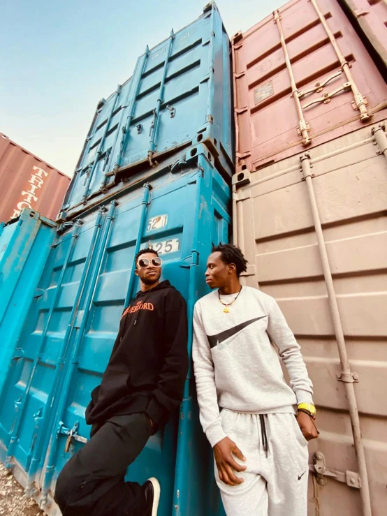 two men standing next to each other in front of shipping containers, an album cover, trending on pexels, afro tech, streetwear, profile image, low angle shot