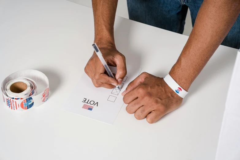 a person writing on a piece of paper next to a cup of coffee, by Joe Bowler, spiked wristbands, election poster, official product photo, 9 9 designs