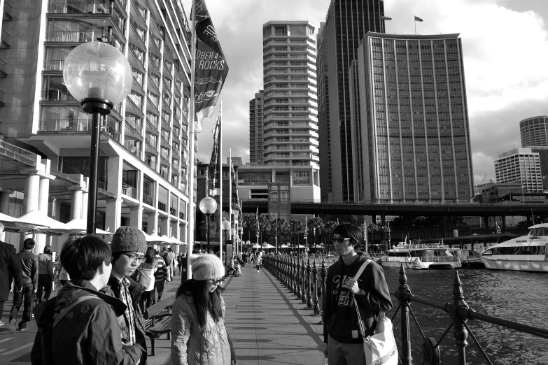 a group of people walking down a sidewalk next to a body of water, a black and white photo, inspired by Thomas Struth, hurufiyya, sydney, lots of building, portrait of tall, high quality upload