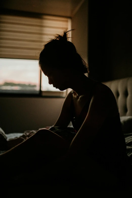 a woman sitting on top of a bed next to a window, inspired by Elsa Bleda, trending on pexels, silhouette of man, her gaze is downcast, underexposed, upset