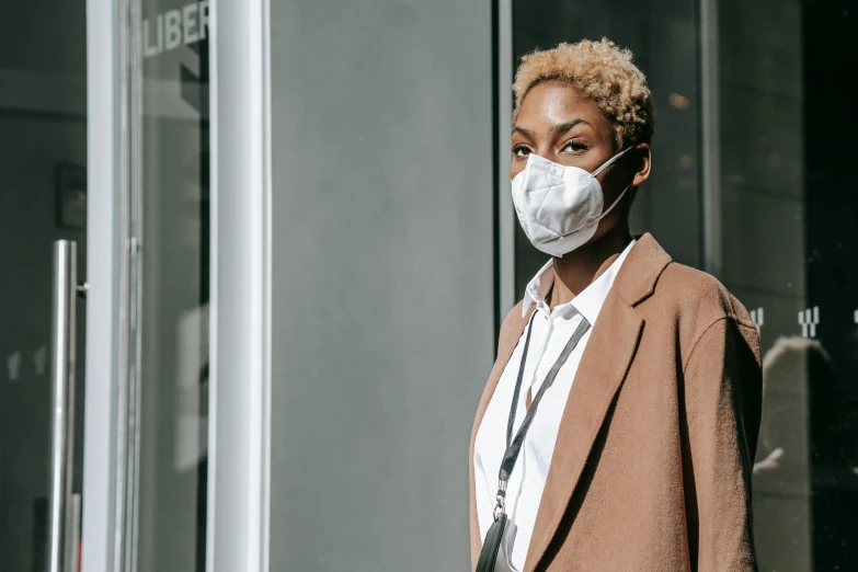 a woman wearing a face mask standing in front of a building, by Emma Andijewska, pexels contest winner, afrofuturism, wearing a labcoat, short blonde afro, exiting store, wearing a brown