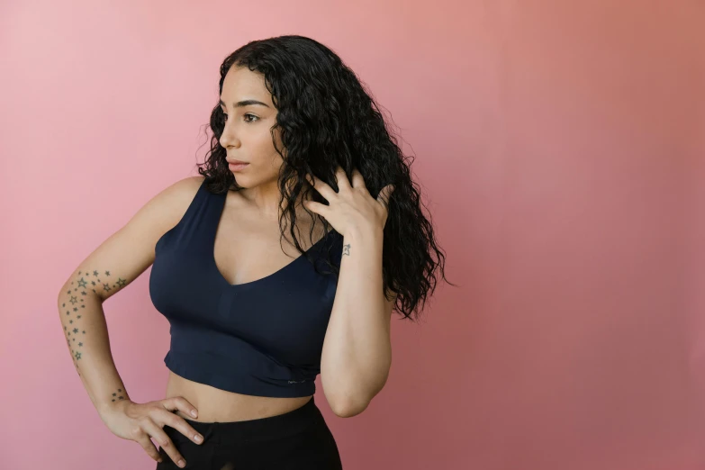 a woman standing in front of a pink wall, inspired by Esaias Boursse, trending on pexels, arabesque, wearing a cropped black tank top, long black curly hair, blue tight tshirt, various posed