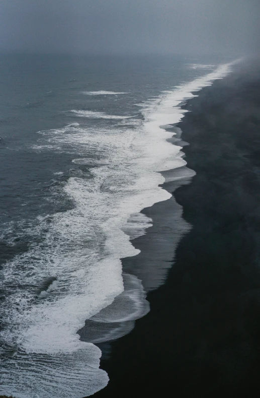 a large body of water next to a beach, inspired by Andreas Gursky, black sand, ocean spray, afar, 4k movie still