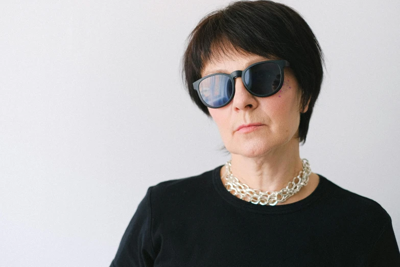 a woman wearing sunglasses standing in front of a white wall, inspired by Cornelia Parker, unsplash, short dark hair, portrait 6 0 - year - old woman, hasbulla magomedov, wearing collar