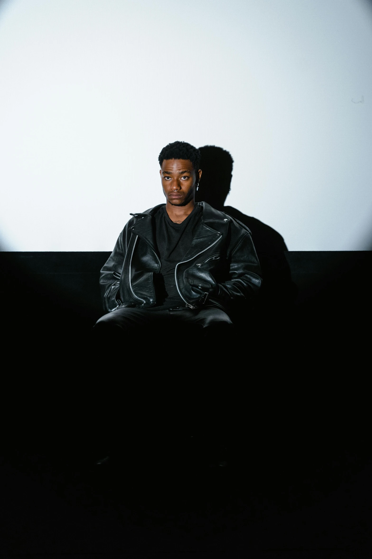 a man sitting on a couch in front of a projector screen, by Cosmo Alexander, wearing a leather jacket, 2 1 savage, black reflect robe, looking towards the camera