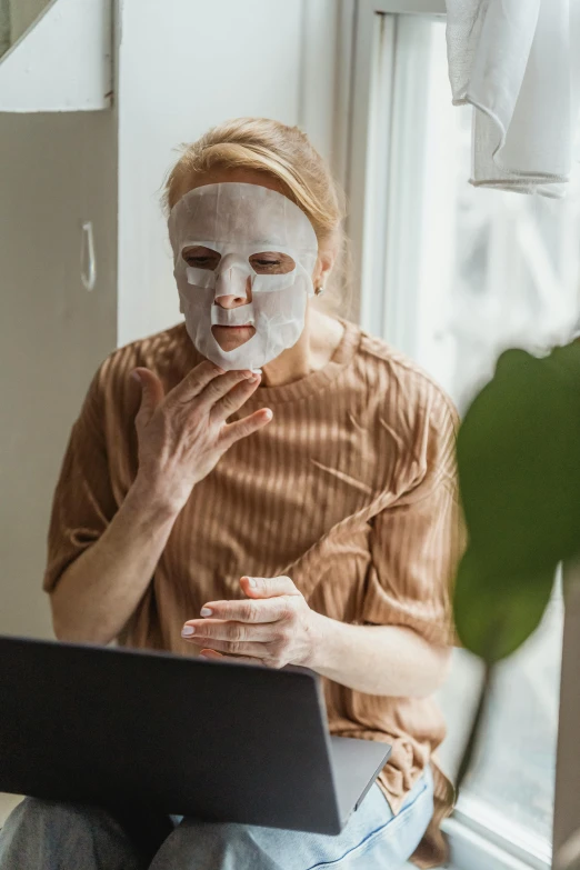 a woman sitting on a window sill using a laptop, a picture, by Julia Pishtar, trending on pexels, renaissance, the mask covers her entire face, white man with black fabric mask, skincare, machine parts embedded into face